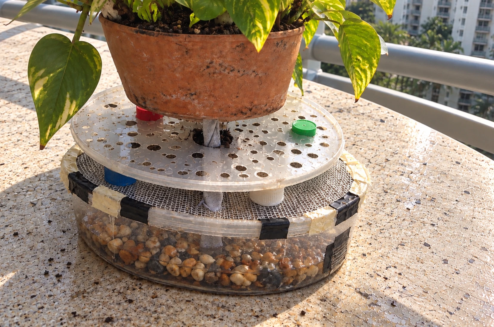 Physical prototype: a clear tray of pebbles under a perforated platform, supporting a terracotta pot on a balcony.